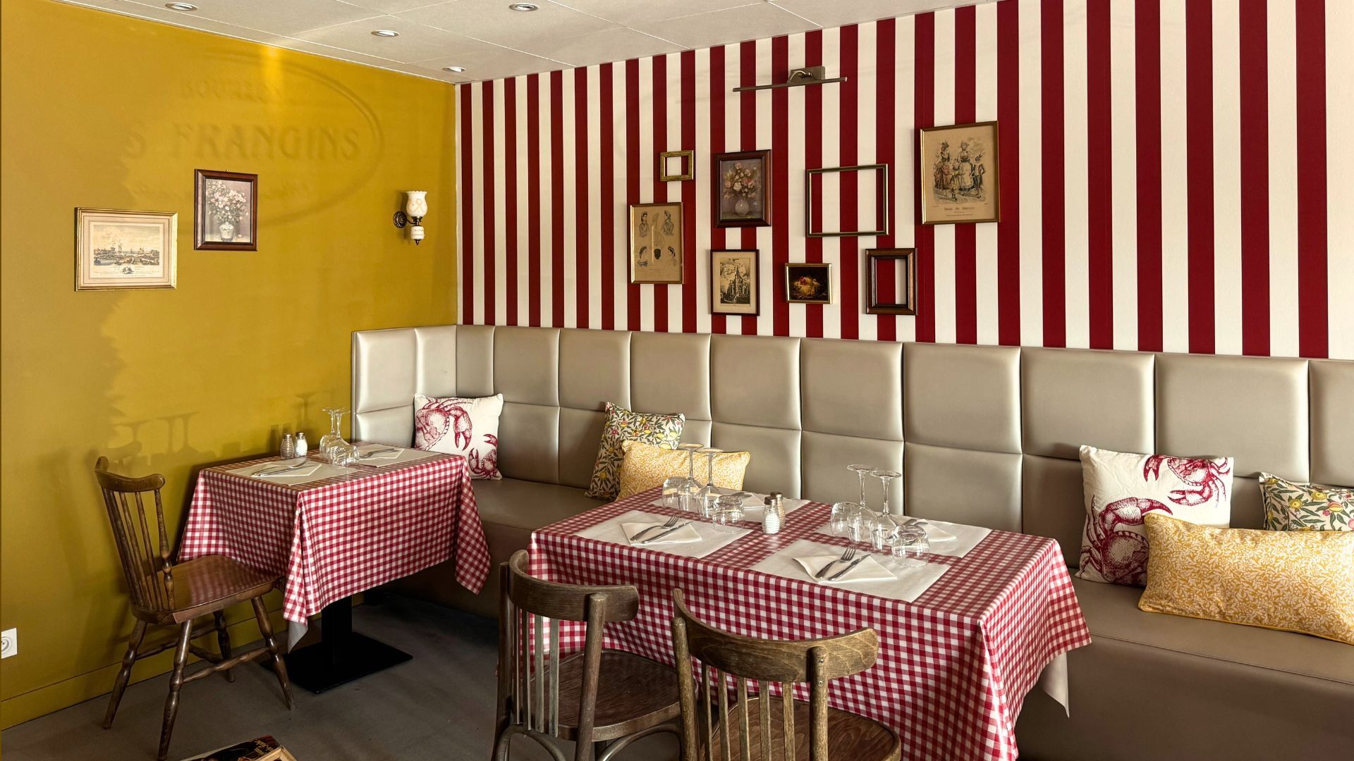 La salle du Bouillon : longue banquette, coussins, nappes vichy rouges, mur rayé rouge et blanc orné de cadres anciens, mur ocre.